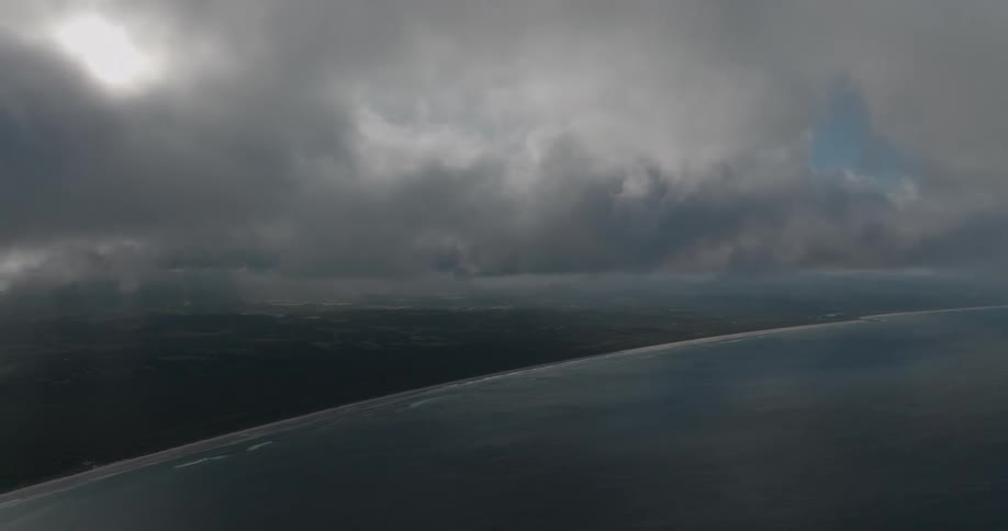 A dramatic aerial view shows the vast ocean meeting the coastline under a sky of dark, moody clouds with the sun breaking through