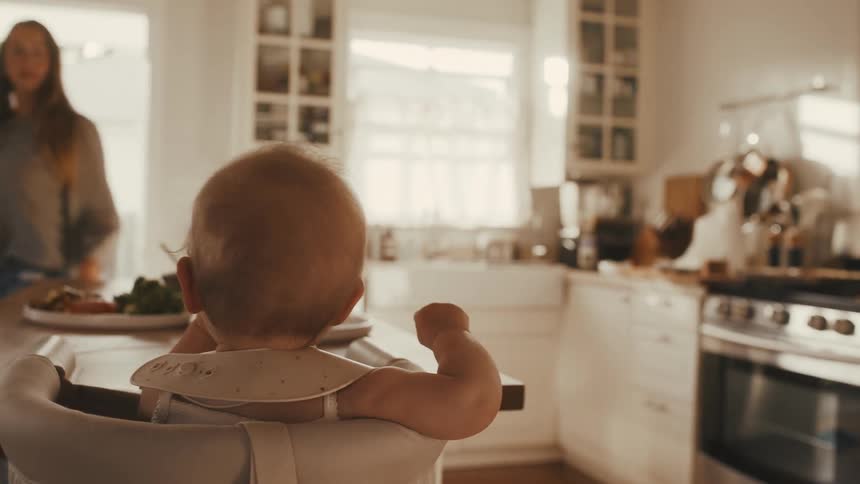 A baby sits patiently in a highchair, watching from behind as its mother happily dances around the sun-drenched kitchen