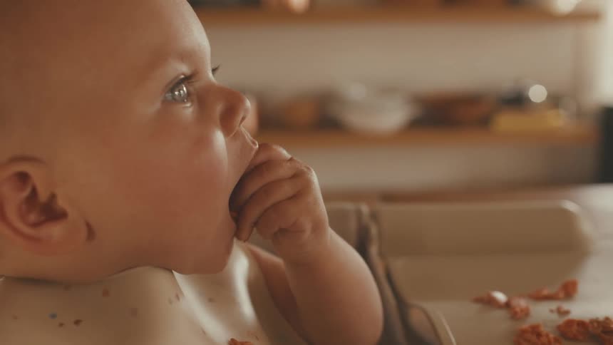 A close-up slow-motion shot of an adorable baby in a high chair learning to eat solid food on its own