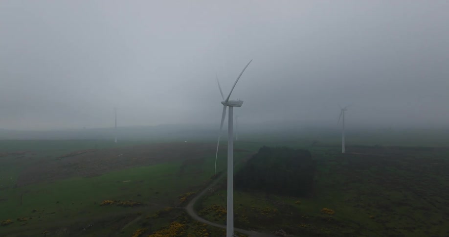 An aerial view of a wind farm shows large turbines slowly spinning on a green hill during a very foggy, overcast day