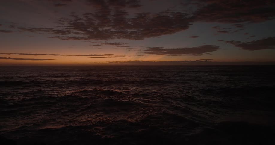 A dark, moody ocean with gentle waves rolling under a cloudy sky as the last light of sunset fades on the horizon