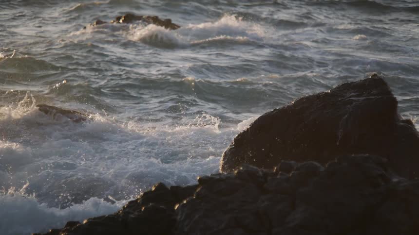 Ocean waves crash against dark, jagged rocks in dramatic slow motion as the golden hour sun glistens on the water