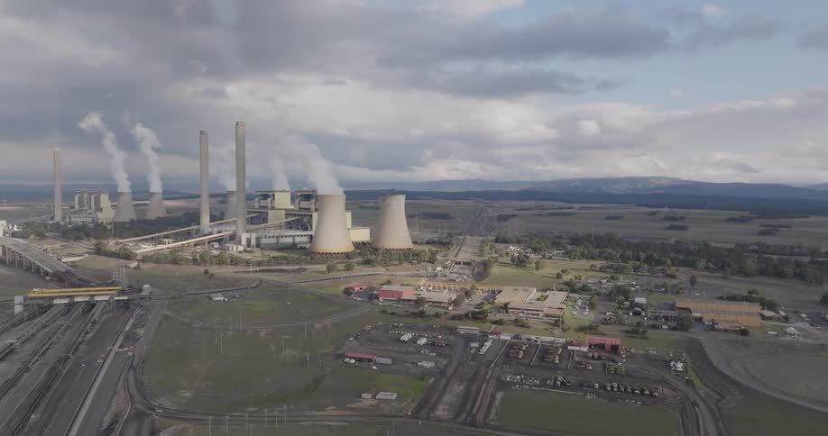 An expansive aerial view captures a large coal power plant, its towering smokestacks and cooling towers releasing steam into the cloudy sky