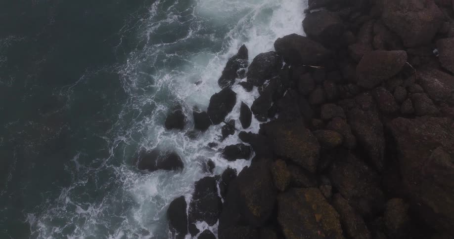 Slowly descending aerial shot of powerful ocean waves crashing against a rugged, rocky coast on a foggy day