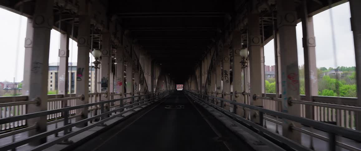 A forward-moving POV shot travels down the center of a long, dark, industrial metal bridge covered in graffiti