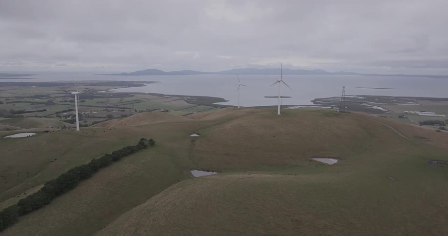 A wide aerial view shows several wind turbines spinning on rolling green hills overlooking the ocean on an overcast day