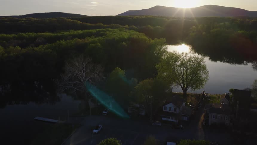 A beautiful aerial dolly shot reveals a tranquil lakeside community and an expansive forest at sunset, with mountains on the horizon