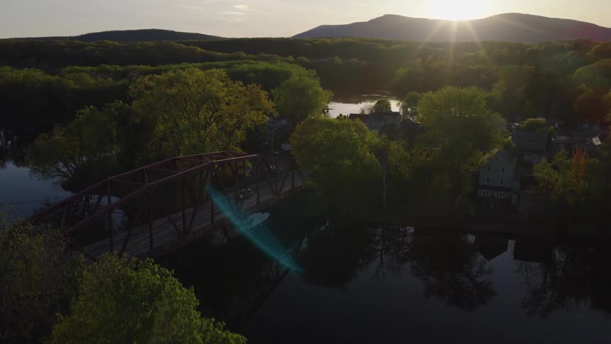 A serene aerial flight over a lush, green landscape towards a distinctive red bridge crossing a river as the sun sets behind distant mountains