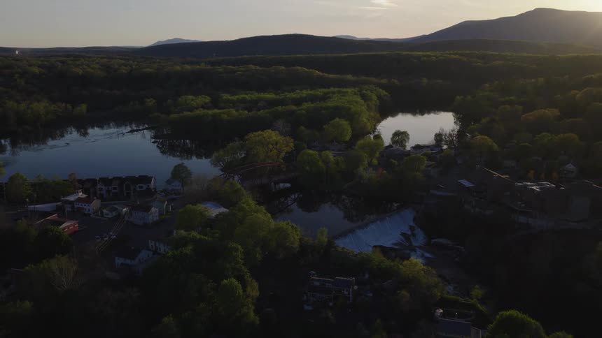 An aerial view pulls back from a quaint small town with a red bridge over a tranquil river at sunset, revealing a scenic waterfall and distant mountains