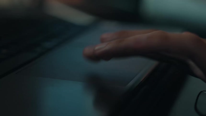 A dramatic close-up of a person's hand using a laptop's trackpad in a dimly lit room