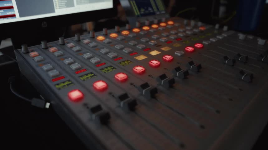 A professional audio mixing console with illuminated lights and faders in a dark recording or broadcast studio