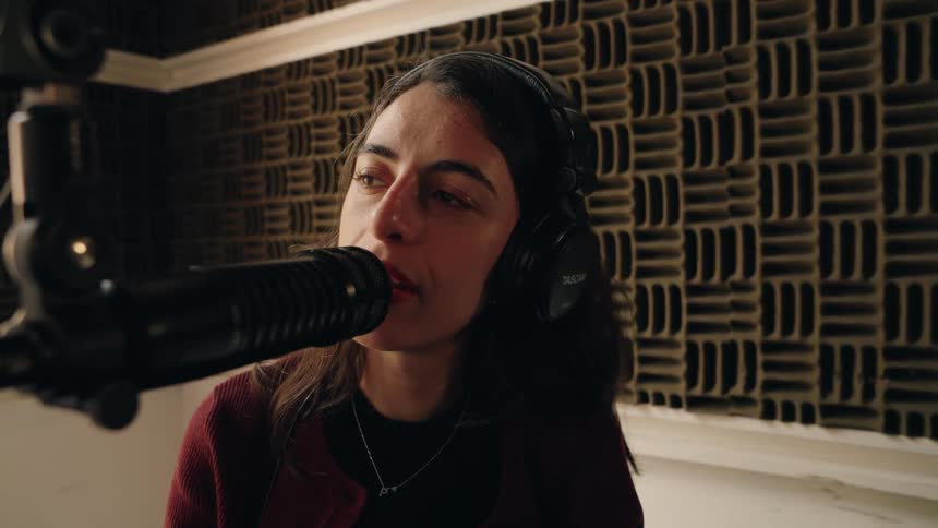 A woman wearing headphones speaks passionately into a microphone while recording in a soundproof studio