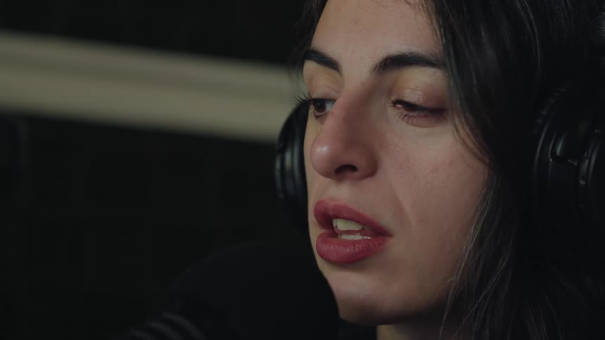 A close-up side profile of a woman with long dark hair speaking into a microphone while wearing headphones in a studio