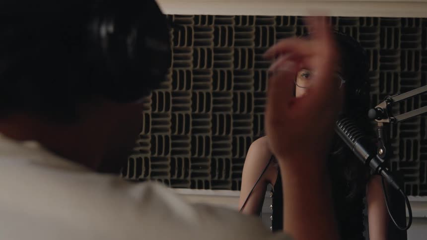 A young woman with glasses and headphones hosts a podcast, smiling as she converses with her male guest in a soundproof recording studio