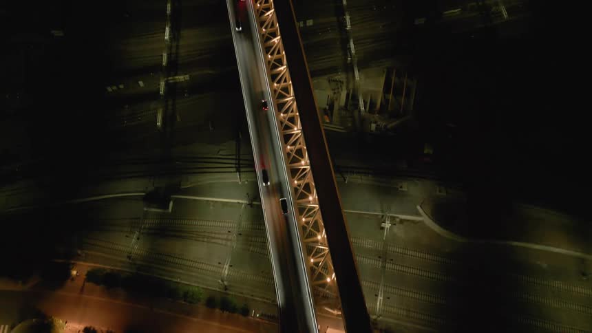 An ascending top-down aerial shot captures cars driving over a beautifully illuminated city bridge and railyard at night
