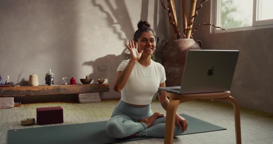 A cheerful woman sitting in a yoga pose waves hello to a laptop screen during a virtual class in a peaceful studio