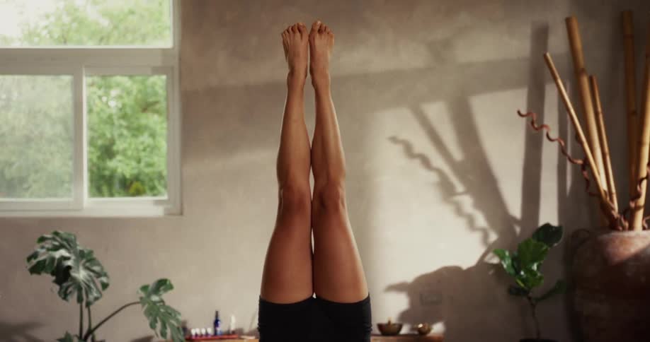 Close up of a woman's legs held steady in a yoga inversion pose inside a serene, sun-dappled studio
