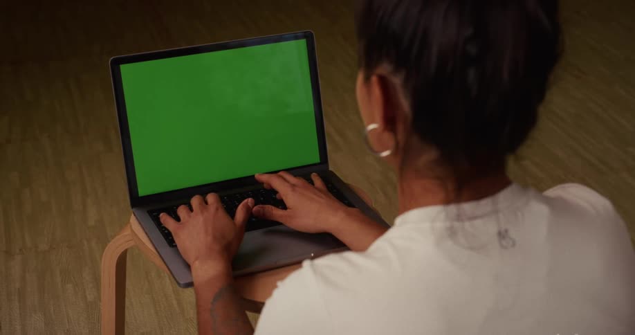 Over-the-shoulder shot of a woman with dark hair typing on a laptop with a blank green screen