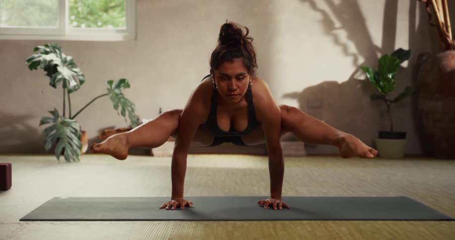 A strong and flexible woman practices an advanced firefly yoga pose in a peaceful, sunlit studio