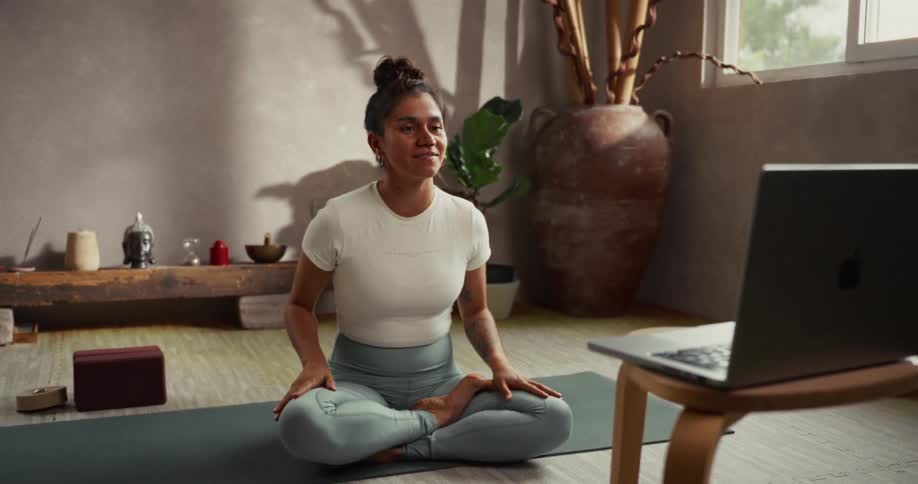 A smiling yoga instructor guides an online class, demonstrating a breathing exercise in her calm studio