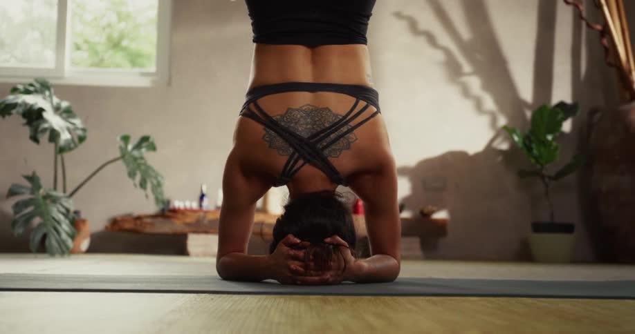 A strong woman with a prominent back tattoo holds a steady headstand on a yoga mat in a serene, sunlit studio