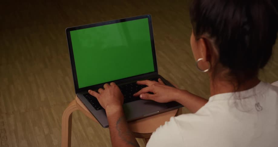 An over-the-shoulder view of a woman with her hair up, typing on a laptop computer with a green screen display