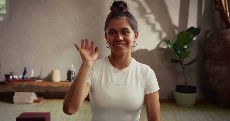A friendly woman with her hair in a bun waves hello while sitting in a serene yoga studio