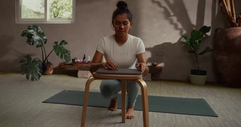 A woman in workout gear carefully places her laptop on a stool in a serene studio, preparing for an online yoga or fitness class