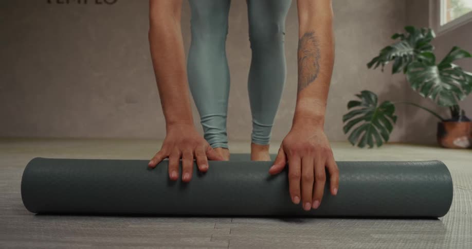 A person with a tattoo on their arm carefully unrolls a yoga mat in slow motion to begin their practice