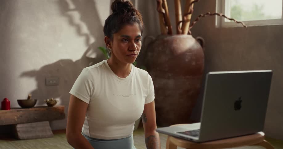 A woman in yoga wear sits on a mat in a serene, sunlit room and begins her online guided meditation by pressing play on her laptop