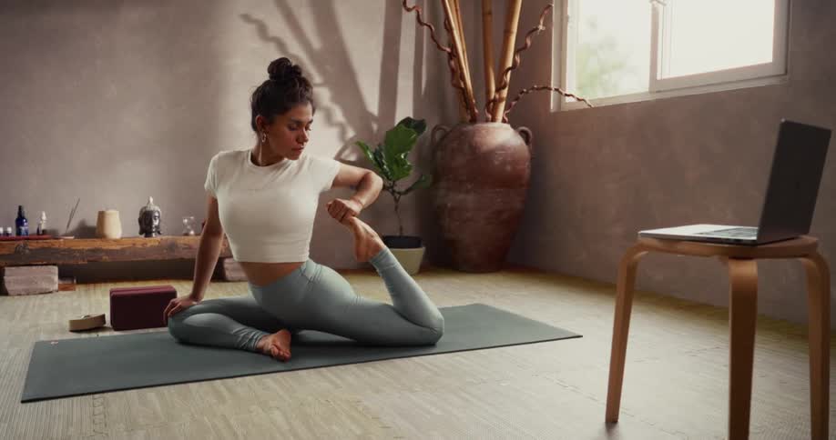 A flexible woman holds a deep pigeon pose stretch on her yoga mat while following an online class on her laptop in a tranquil, sunlit studio