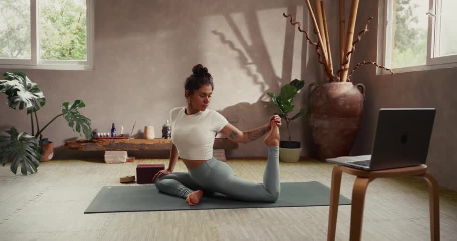 A flexible woman holds a deep yoga stretch on a mat in a serene, sunlit studio while following an online class on her laptop