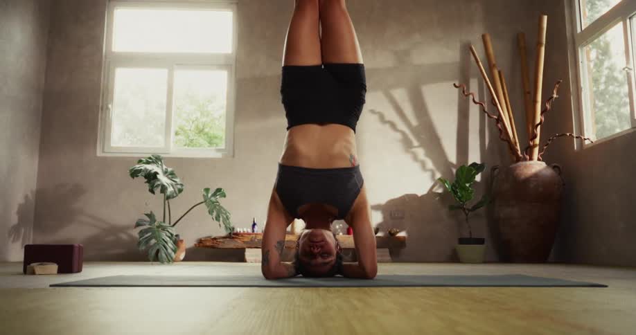 A serene woman with her eyes closed maintains a perfect headstand yoga pose in a sunlit, peaceful studio