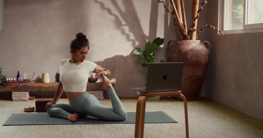 A woman follows an online yoga class on her laptop, holding a deep stretching pose in a tranquil, sunlit studio