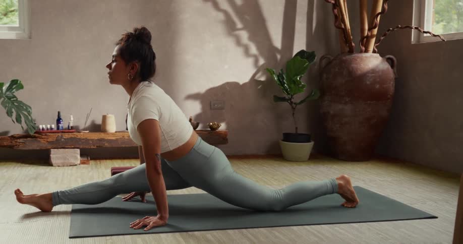 A woman practicing yoga at home follows a virtual class on her laptop, smoothly transitioning into a full split pose on her mat