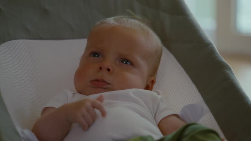 A sleepy newborn baby with blond hair and blue eyes slowly wakes up in a baby bouncer