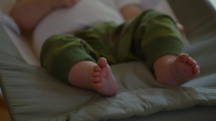 A peaceful newborn baby wearing green pants rests in a cozy bouncer, with a focus on their tiny bare feet