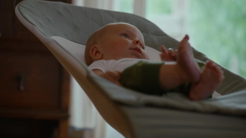 A curious and content newborn baby with blue eyes rests peacefully in a bouncer chair indoors