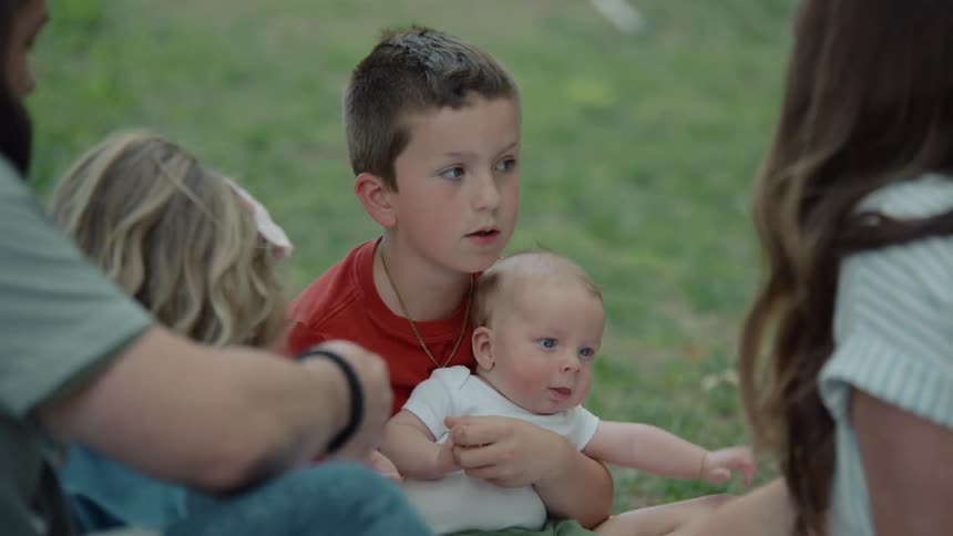 A young boy gently holds his newborn baby brother while sitting on the grass with his family