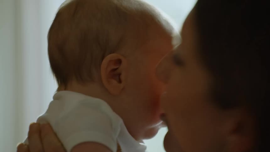 An affectionate mother gently kisses her newborn baby's cheek in a tender, close-up shot