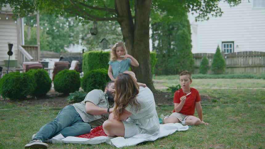 A family of five relaxes on a picnic blanket in their backyard while the children blow bubbles