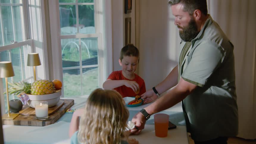 A bearded father serves his son and daughter their meal at a sunlit dining room table