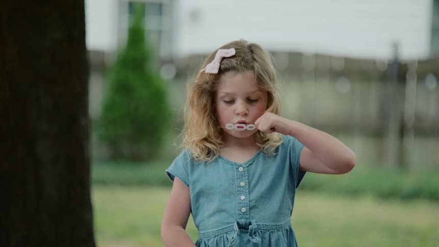 A young girl with blonde hair and a pink bow in her hair blows bubbles in her backyard in slow motion