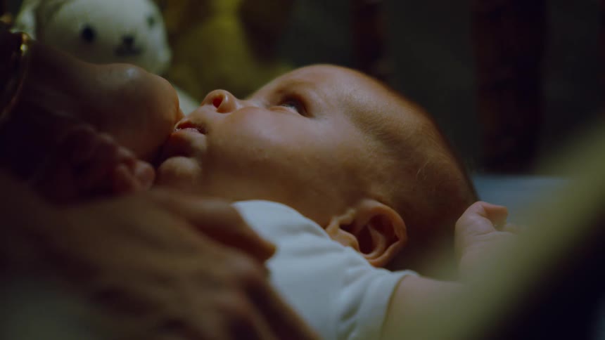 An intimate close-up shows a newborn baby yawning and nursing while being held by its mother in a dark room