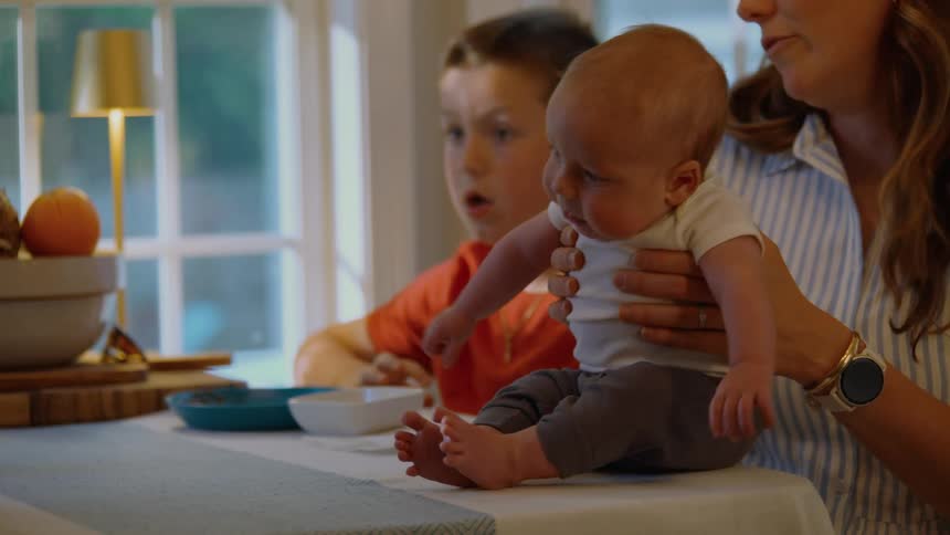 A mother holds her newborn baby on the table as her older son looks on, all captivated by something off-screen