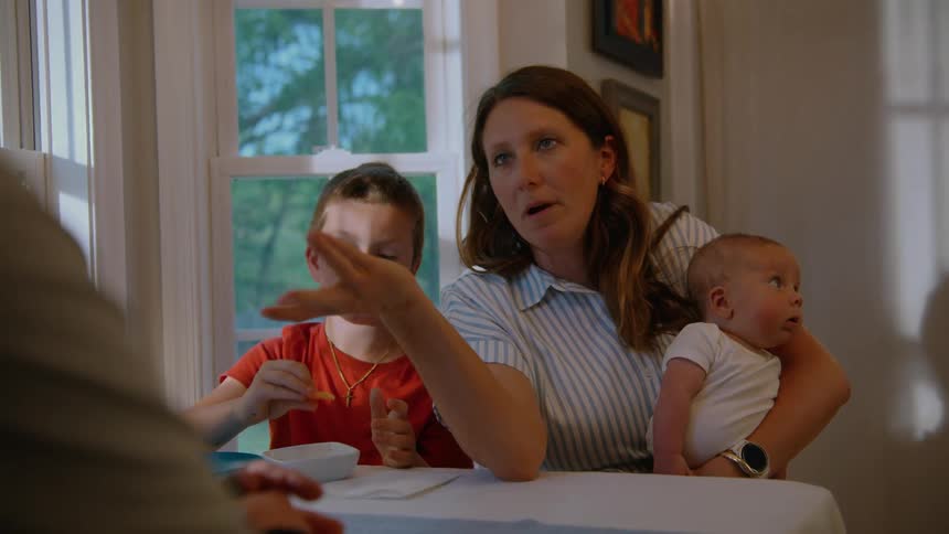A tired mother holding her newborn baby has a conversation with her family at the dinner table