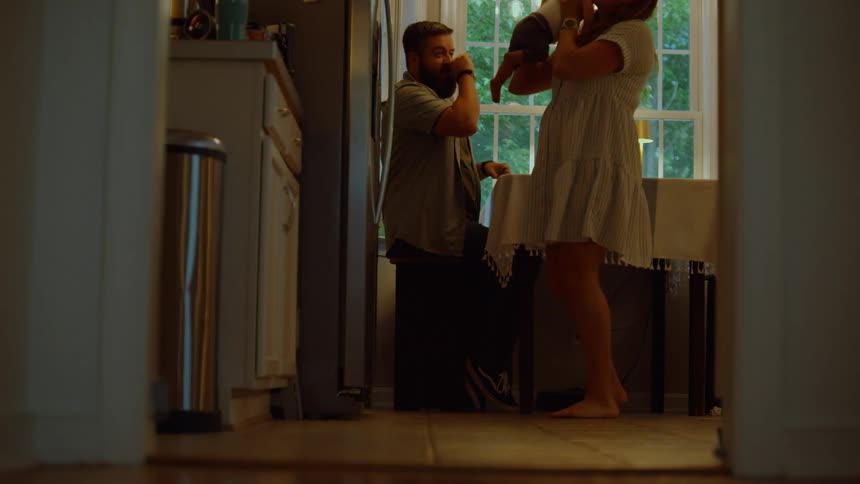 A mother walks into the kitchen and takes her baby from the father who is sitting at the table