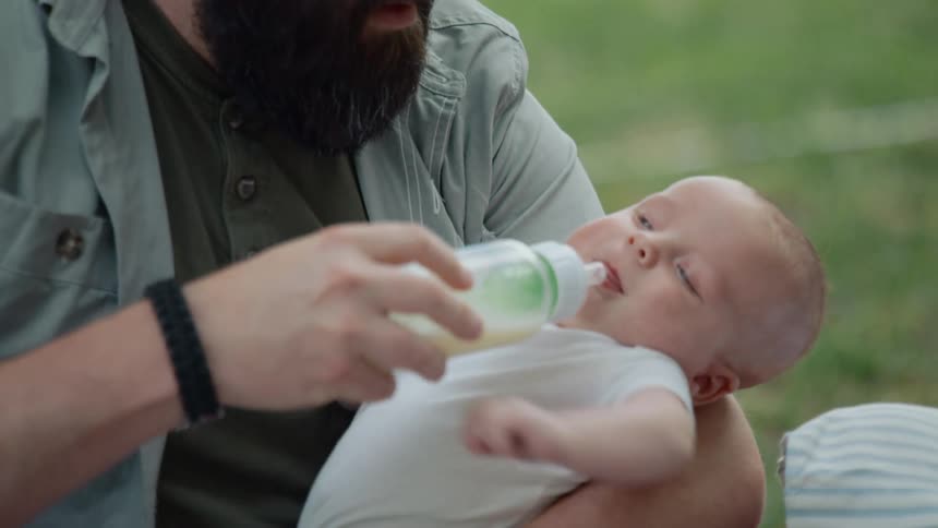 A mother hands her newborn baby to the father, who then lovingly feeds him from a bottle while cradling him