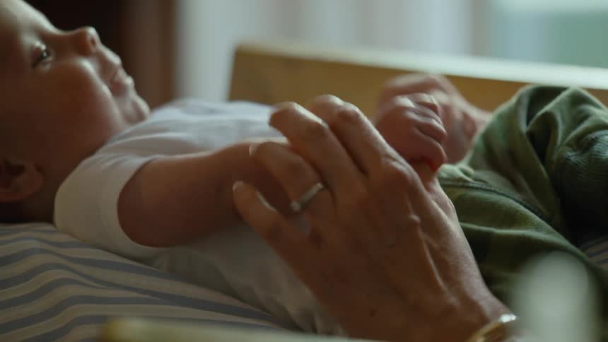 A mother gently holds her infant's hands in a tender moment as the baby lies peacefully and looks up with a smile