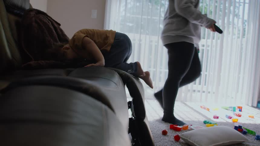 A determined toddler with curly hair successfully climbs up onto the living room couch while his mother is in the background